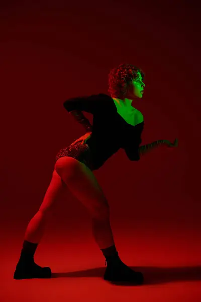 A young, curvy redhead woman in a bodysuit stands confidently in front of a vivid red background. — стоковое фото