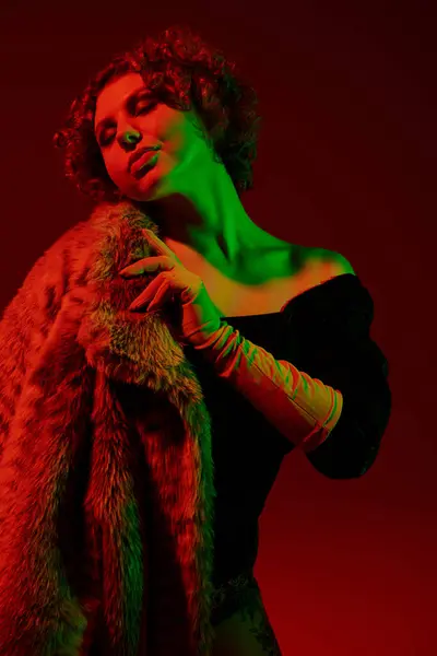 A young, curvy redhead woman strikes a pose in a fur coat against a bold red backdrop. — Stockfoto