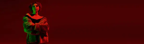 A young, curvy redhead woman in a fur coat stands boldly in front of a rich red background. — Stock Photo