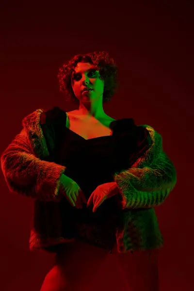A young curvy redhead woman in a fur coat striking a pose in front of a vibrant red light. — Stock Photo