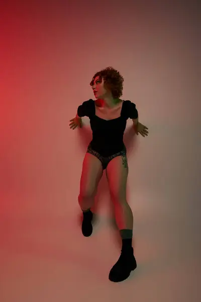 A young curvy redhead woman in a bodysuit dancing energetically in front of a striking red background. — Stock Photo