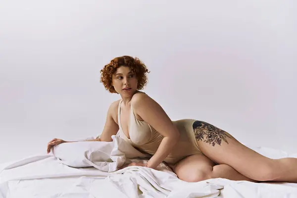 Young, curvy redhead woman with tattoos laying on bed in lingerie against grey background. — стоковое фото