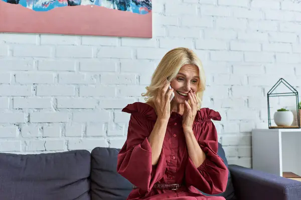 Mature woman in stylish dress, seated on couch, deeply engaged in cell phone conversation. — Stock Photo