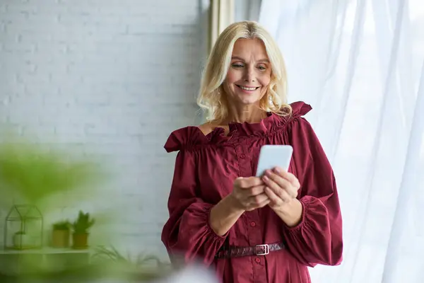 A sophisticated woman in stylish dress standing by a window, focusing on her cell phone. — Stock Photo
