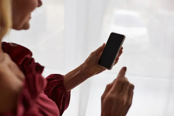 Mature woman in a red shirt stylishly holds a cell phone at home. — Stock Photo