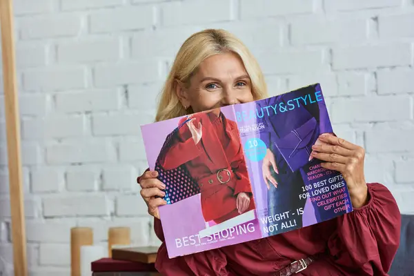 A mature woman in a stylish dress holds a magazine in front of her face. — Stock Photo