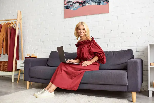 Eine reife Frau in einem stylischen Kleid sitzt auf einer Couch und konzentriert auf ihrem Laptop. — Stockfoto