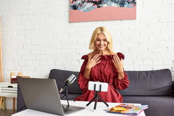 Mujer madura en vestido rojo chic creando un podcast sobre la belleza femenina delante de un ordenador portátil. - foto de stock