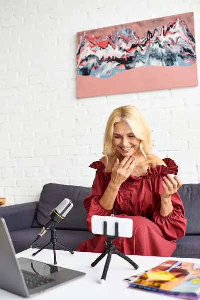 A mature elegant woman in a red dress is sitting at a table in front of a laptop computer, recording a podcast on female beauty. — Stock Photo
