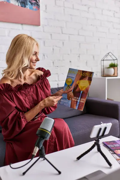 Una donna matura ed elegante vestita di rosso si siede su un divano, assorbita dalla lettura di una rivista e dalla creazione di podcast. — Foto stock