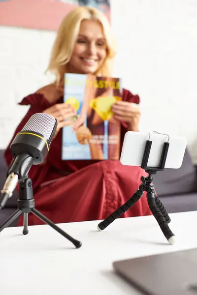 Una donna matura ed elegante in un abito rosso chic seduta davanti a un microfono, con in mano una rivista. — Foto stock
