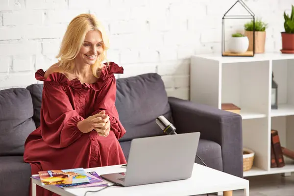 Maduro elegante mujer en rojo elegante vestido podcasting en belleza femenina utilizando el ordenador portátil en el sofá. - foto de stock