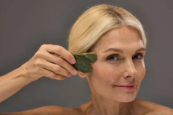 Mature woman using gua sha on a gray backdrop. — Stock Photo