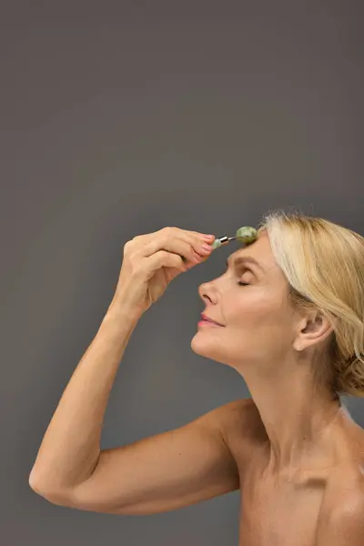 Beautiful mature woman using face roller on a gray backdrop. — Stock Photo