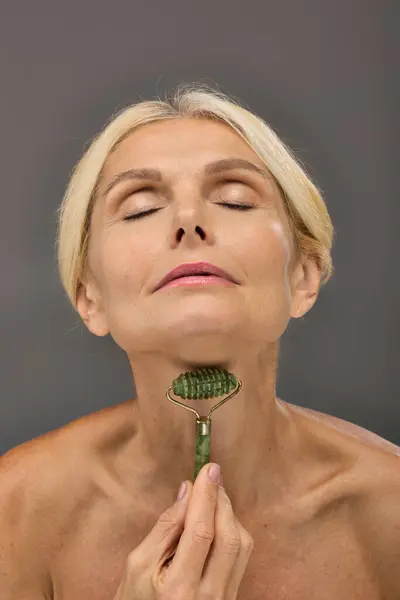 Lovely mature woman using face roller on a gray backdrop. — Stock Photo