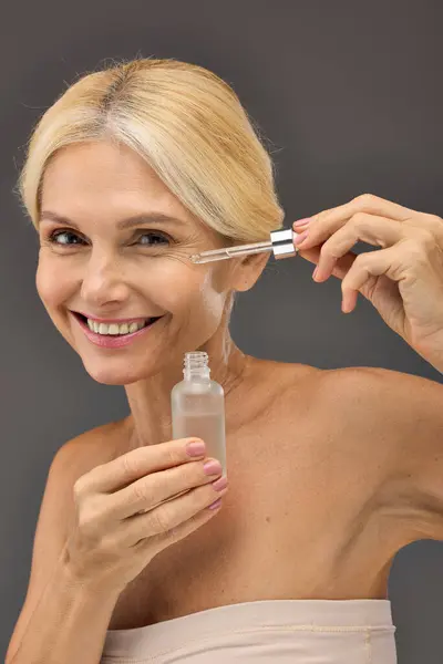 Good looking mature woman applying serum on a gray backdrop. — Stock Photo