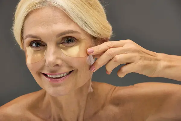 Mature woman showcasing eye patches while applying skincare. — Stock Photo