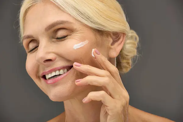 Una donna matura applica gioiosamente la crema per la cura della pelle sul suo viso su uno sfondo grigio. — Foto stock