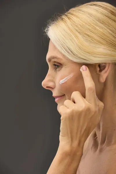 Mulher graciosamente aplicando creme em seu rosto contra um pano de fundo cinza. — Fotografia de Stock