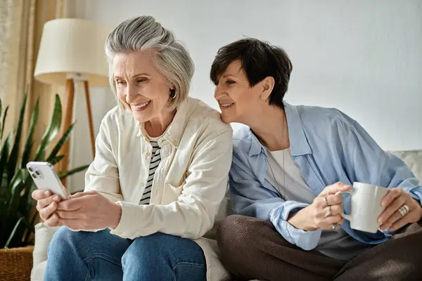Two mature women, a loving lesbian couple, share a moment on a couch, engrossed in a phone. — Stock Photo