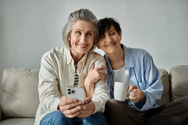 Deux femmes, un couple de lesbiennes mûres et aimantes, assises sur un canapé, absorbées dans un téléphone portable. — Photo de stock