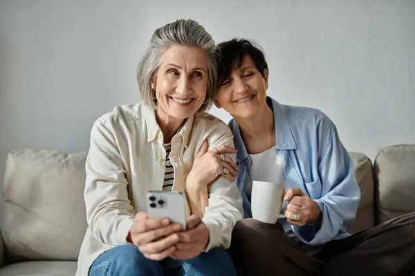 Two women, a loving mature lesbian couple, sit on a couch, engrossed in a cell phone. — Stock Photo