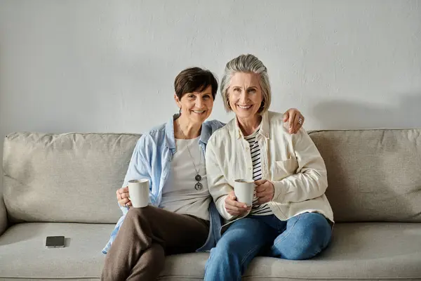 Two senior women relax on a couch, sipping coffee from mugs. — Stock Photo