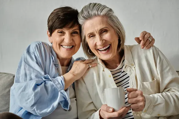 Two older women, a loving mature lesbian couple, sit together on a couch, enjoying a cup of coffee. — Stock Photo