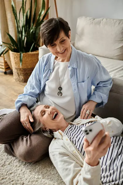 Two women laugh while seated on the floor, engrossed in a cell phone. — Stock Photo