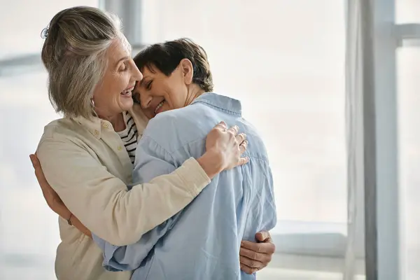 Dos ancianas se abrazan tiernamente a la luz del sol por una ventana. - foto de stock