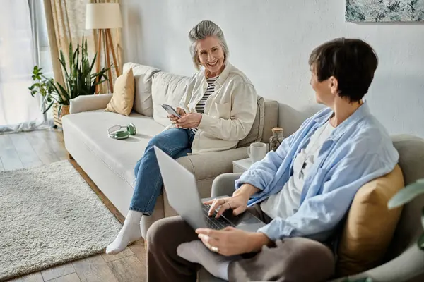 Two people, a loving mature lesbian couple, sit on a couch and use a laptop. — Stock Photo