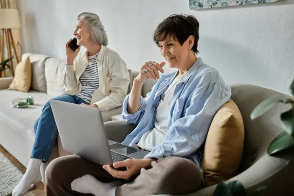 Two women, a loving mature lesbian couple, talking on the phone and using a laptop on a couch. — Stock Photo