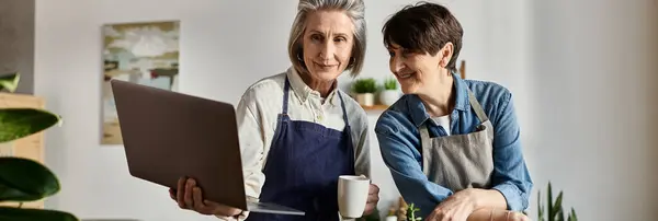 Two women in aprons studying a laptop screen together. — Stock Photo