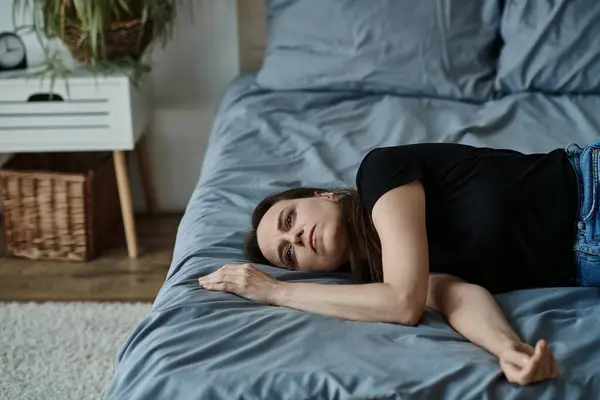 A middle-aged woman lies on her bed with arms crossed, lost in thought. — стоковое фото
