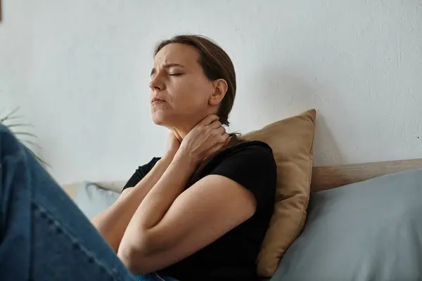 Middle-aged woman sits on bed, hands on neck. — Stock Photo