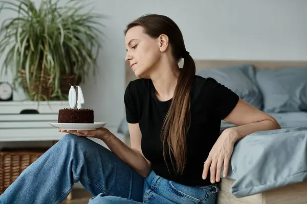 A middle-aged woman sits on a bed, holding a birthday cake. — Stockfoto