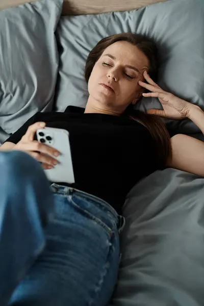Woman lays in bed with phone on head. - foto de stock