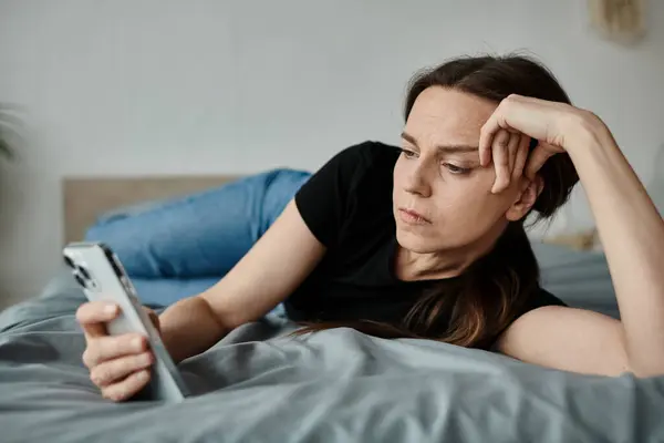 Middle-aged woman laying on bed, absorbed in her phone screen. — Stockfoto