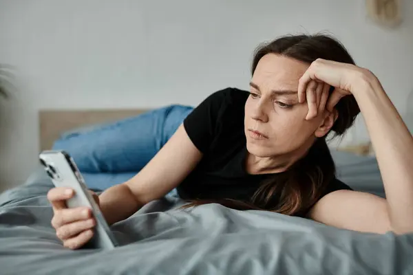 Middle-aged woman laying on bed, absorbed in her phone screen. — стокове фото