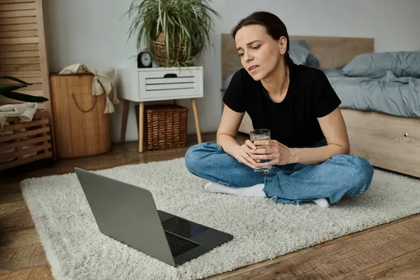 A middle-aged woman sits on the floor with a laptop and a glass of water. — Foto stock
