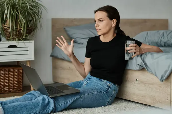 Middle-aged woman sits with laptop and water, absorbed in online therapy session. — стоковое фото