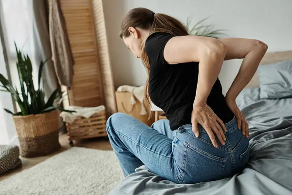 A middle-aged woman sits on a bed, experiencing back pain, struggling with depression. — Foto stock