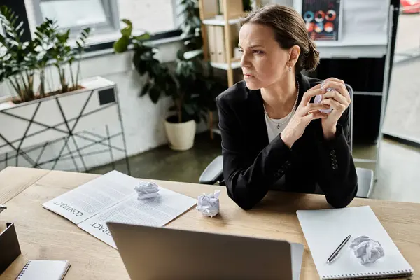 A middle-aged woman sits at a desk surrounded by crumpled paper, showing signs of stress. — Foto stock
