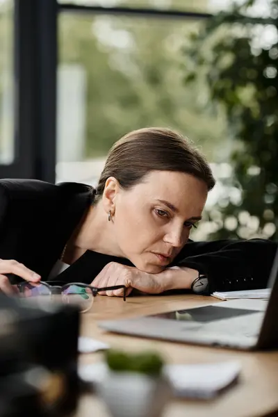 Middle-aged woman experiencing stress is hunched over her laptop in an office setting. — Fotografia de Stock