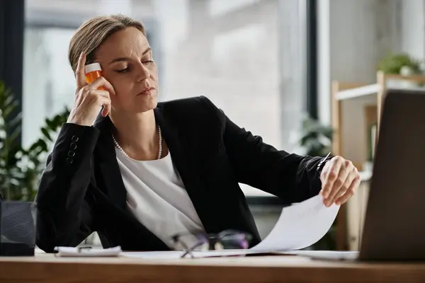 Middle-aged woman sitting at desk, holding bottle of pills. - foto de stock