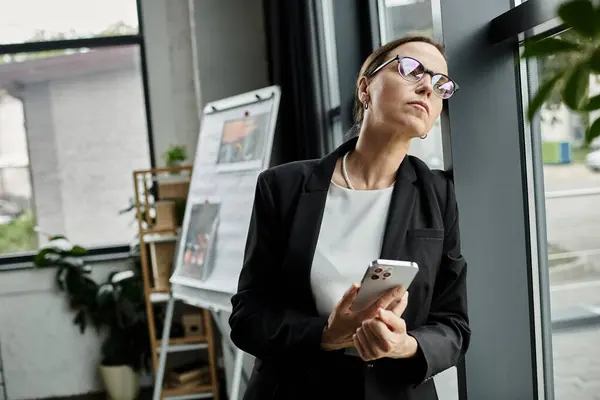 A middle-aged businesswoman looks worriedly at her phone by a window. — стокове фото