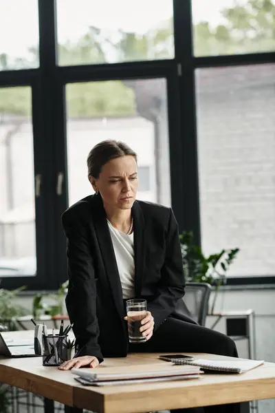 A middle-aged woman appears stressed, sitting at a desk with glass of water. — Fotografia de Stock