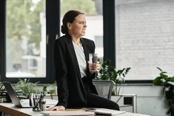 A middle-aged woman in distress sits at a desk, holding a glass of water. — Foto stock