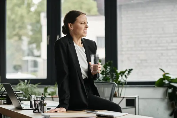 A middle-aged woman in distress sits at a desk, holding a glass of water. — стоковое фото