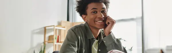 A young man, African American, sitting at a desk with a book, studying online at home. — Stock Photo